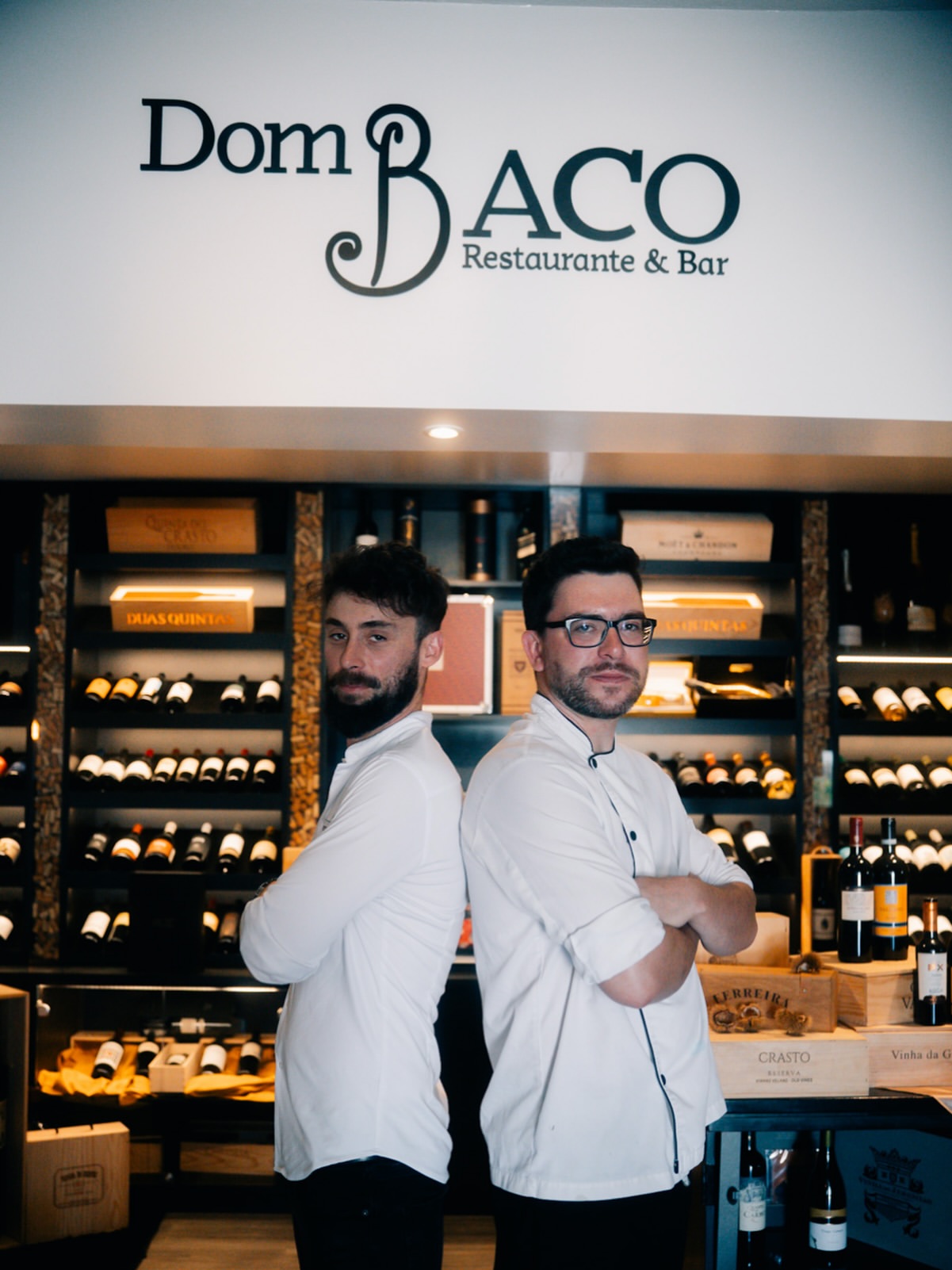 Os dois fundadores do Dom Baco, de costas um para o outro, em frente à adega e ao logotipo do restaurante