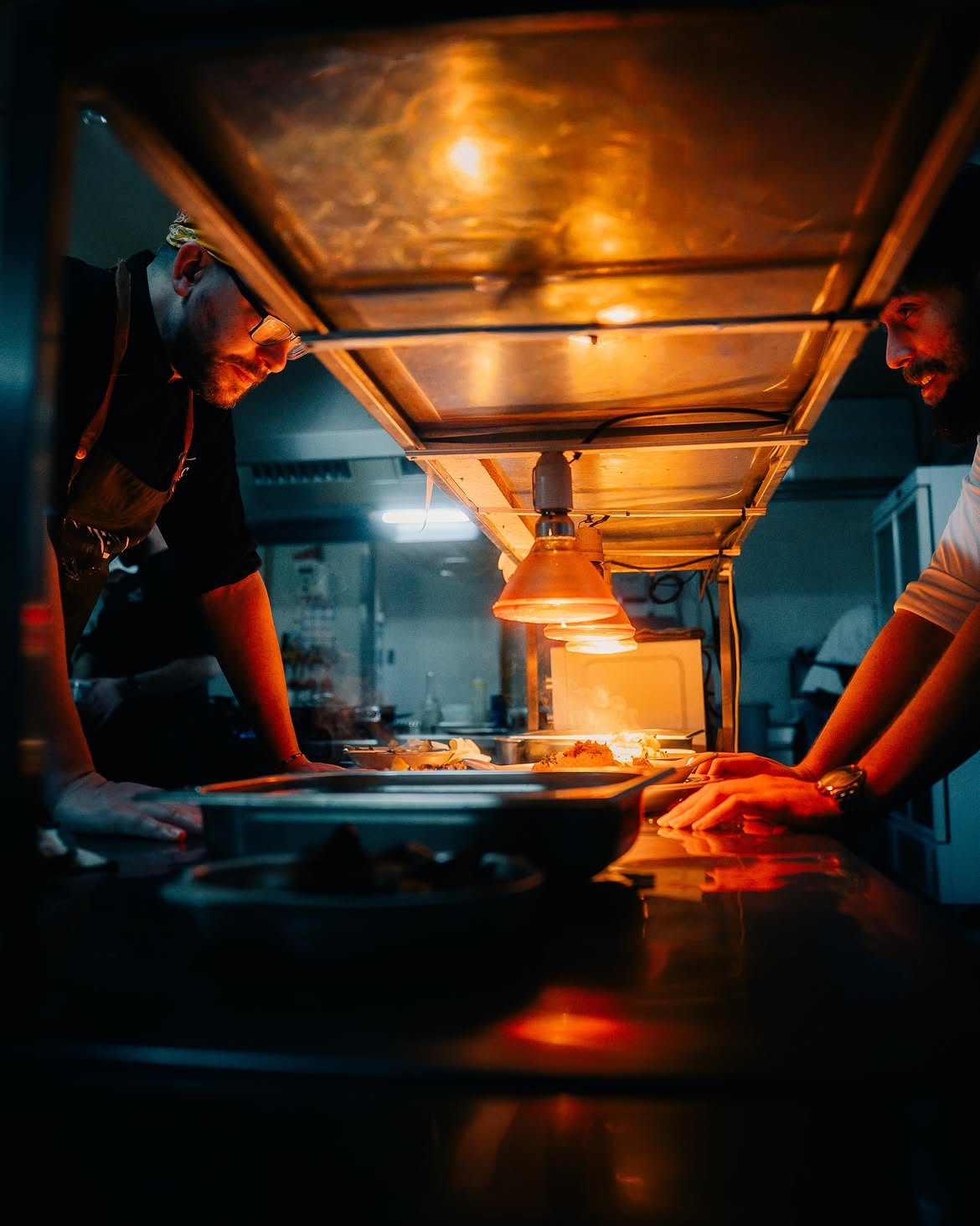 Chef e maître frente a frente no passe da cozinha, sob luz aconchegante